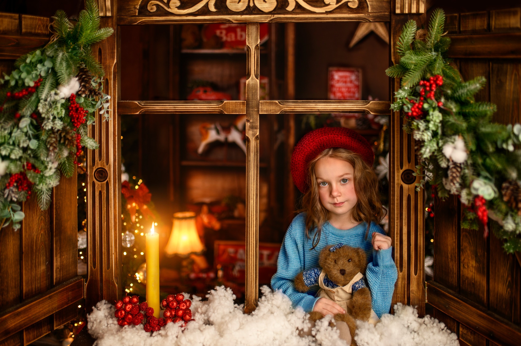 Dziewczynka w czerwonym berecie, trzymająca pluszowego misia, pozuje w drewnianej ramie okiennej udekorowanej świątecznymi gałązkami i świecą, z ciepłym oświetleniem w tle.