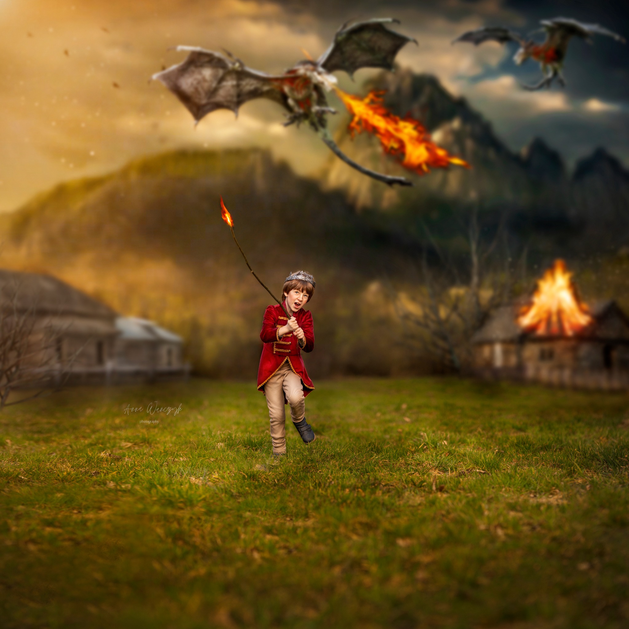 Chłopiec w kostiumie z koroną i płonącą pochodnią ucieka przed ziejącymi ogniem smokami na tle palącej się chaty i górskiego krajobrazu.