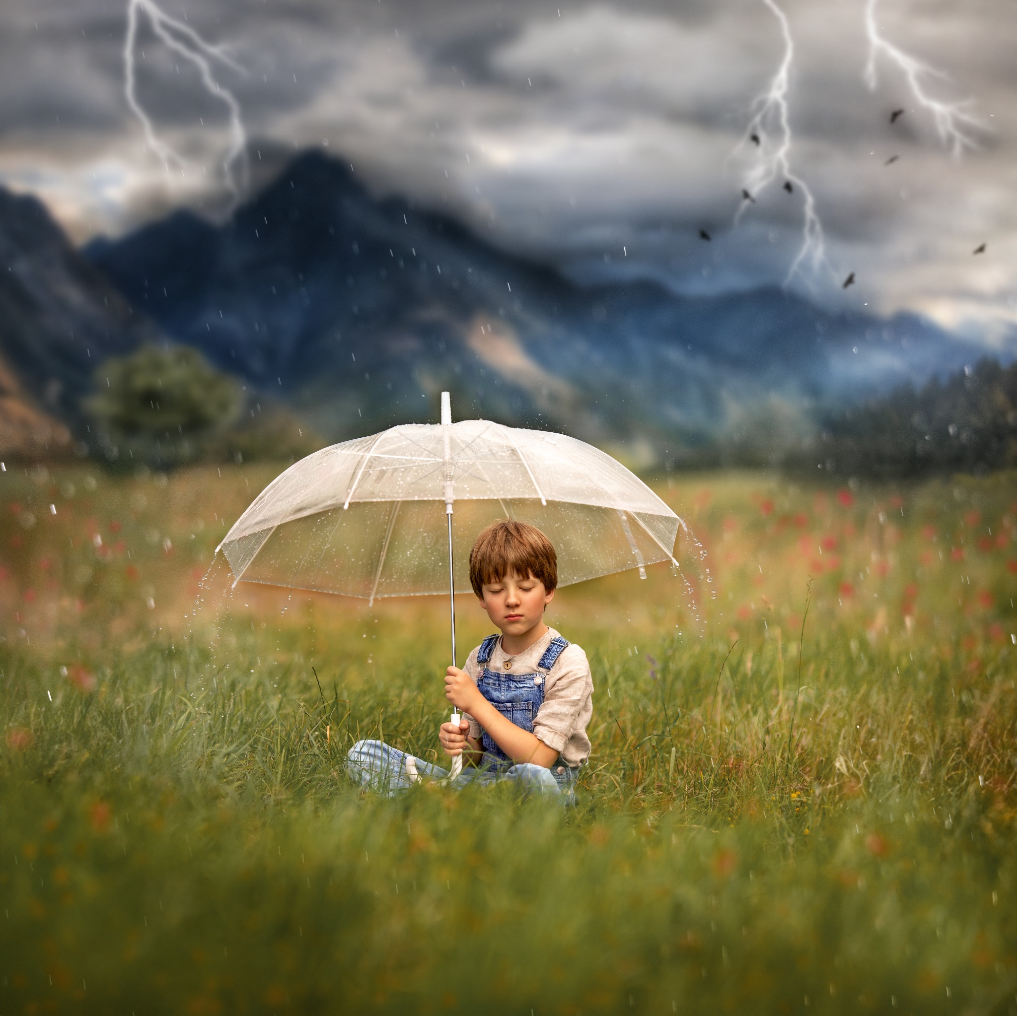 Chłopiec w dżinsowym ogrodniczce siedzi na łące pod przezroczystym parasolem podczas burzy z piorunami, góry w tle, padający deszcz.
