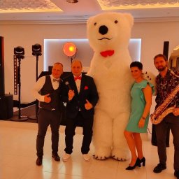 FotoCool Marta Gmyr - Zespół muzyczny z saksofonistą i animatorem w kostiumie białego misia z czerwoną muchą pozuje do zdjęcia na tle dekoracji eventowej z oświetleniem.