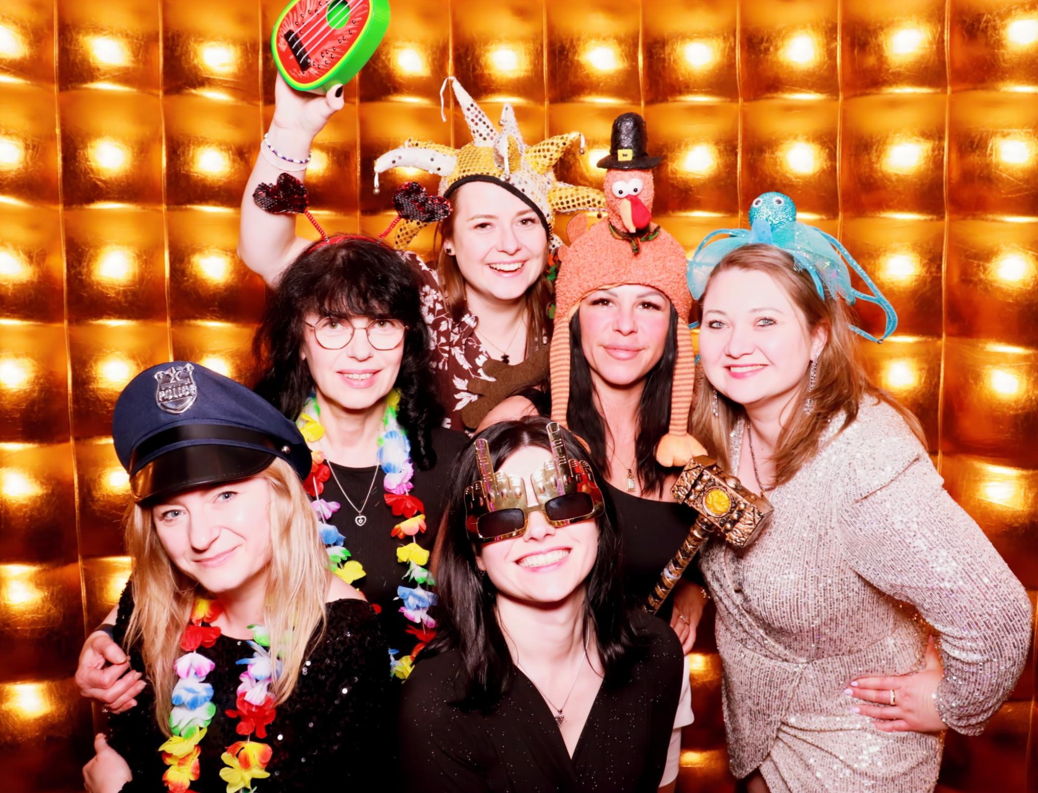 Grupa uśmiechniętych kobiet w przebraniach pozuje na tle złotej ściany, trzymając rekwizyty: kapelusz policjanta, okulary, zabawkowy młotek i inne.