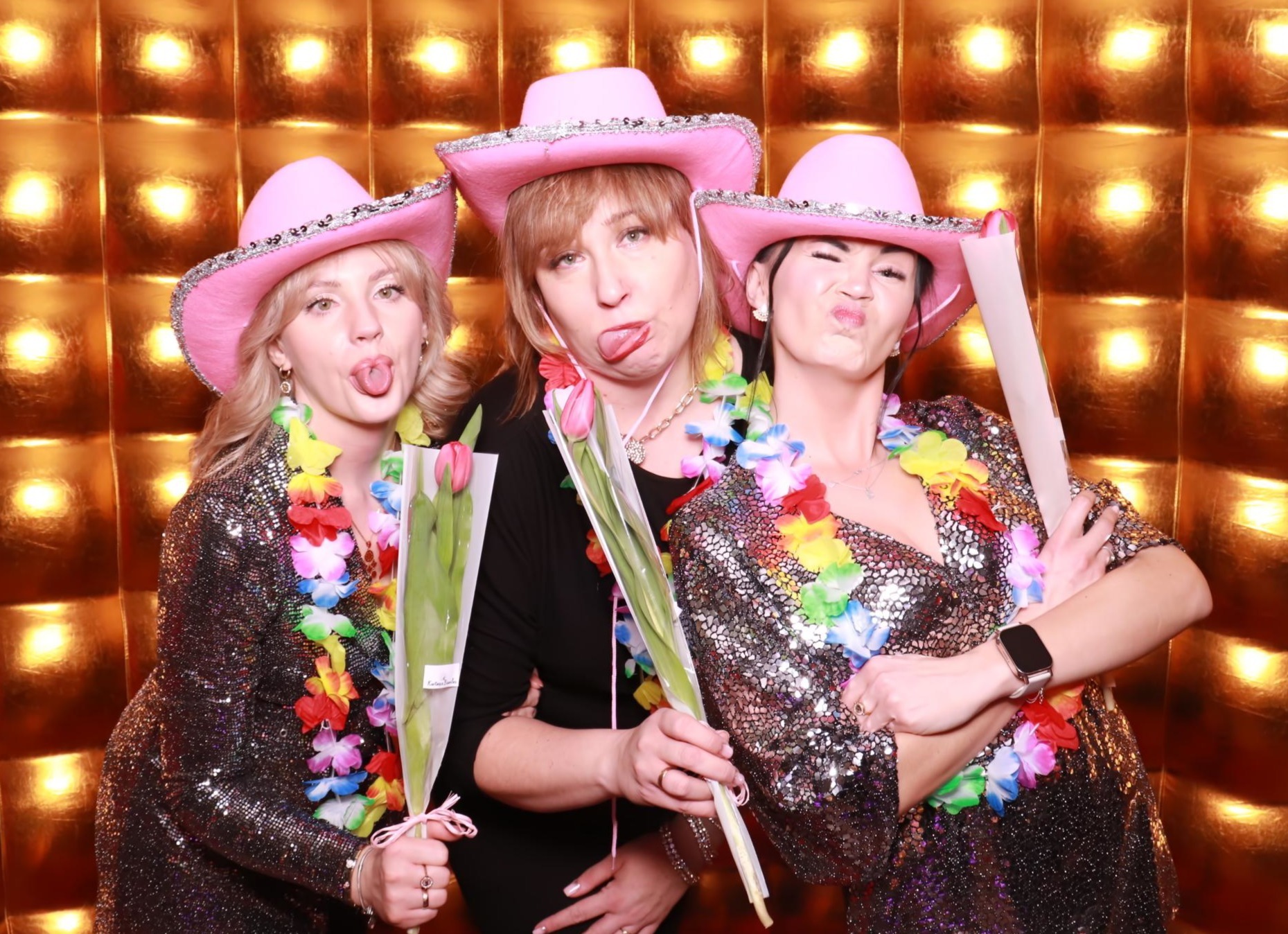 Trzy kobiety w różowych kapeluszach i błyszczących strojach pozują w fotobudce w Płużnicy. Kwiaty i hawajskie naszyjniki dodają koloru. Złote tło.