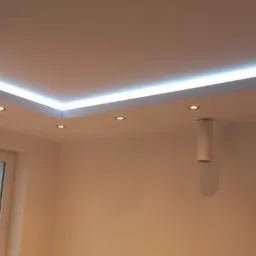 Nowoczesny, biały sufit z oświetleniem LED wzdłuż krawędzi i punktowymi lampami, widok z dołu na jasne wnętrze z oknem.