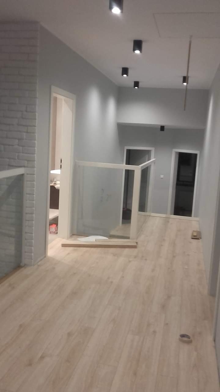 Korytarz w trakcie remontu: świeżo położona jasna podłoga, szare ściany, białe drzwi i balustrada ze szkła, widoczne lampy sufitowe i fragment ściany z cegły.
