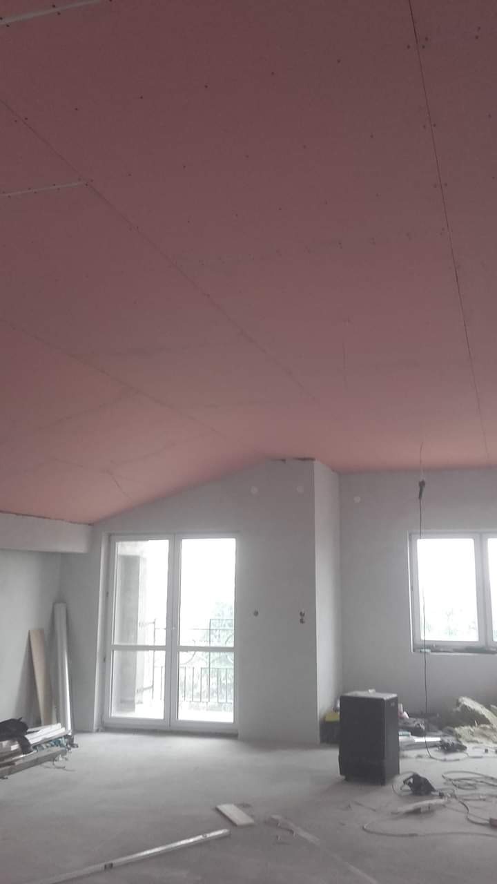 Skośny sufit poddasza pokryty różową płytą gipsowo-kartonową, widoczne łączenia płyt, w tle okna balkonowe i narzędzia budowlane na podłodze, w trakcie prac wykończeniowych.