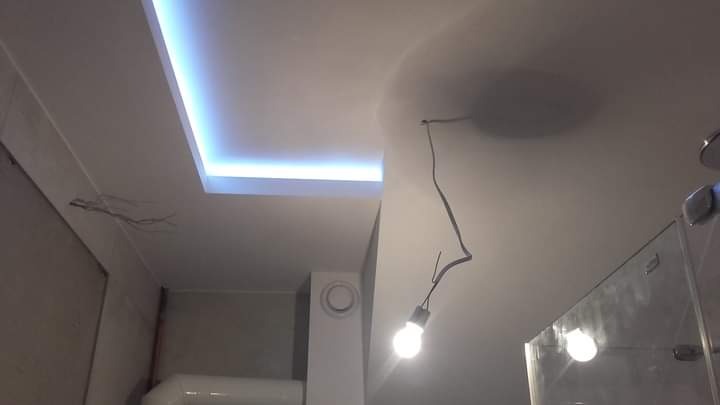 Biały sufit z podwieszanym prostokątnym elementem z niebieskim podświetleniem LED, widoczne kable, żarówka na kablu oraz fragment kabiny prysznicowej w łazience.