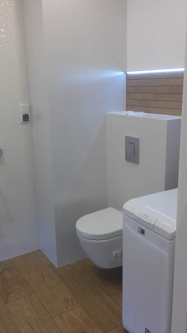 Nowoczesna toaleta w jasnych barwach z drewnianymi akcentami i podświetleniem LED, obok pralka automatyczna.