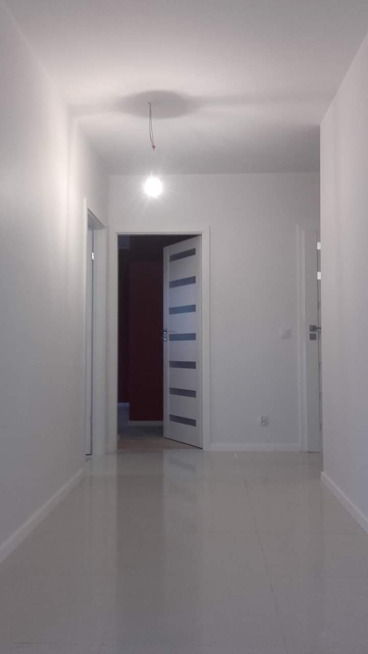 Wnętrze korytarza po remoncie, świeżo pomalowane ściany na biało, połyskująca podłoga z płytek, otwarte drzwi z przeszkleniami w tle.