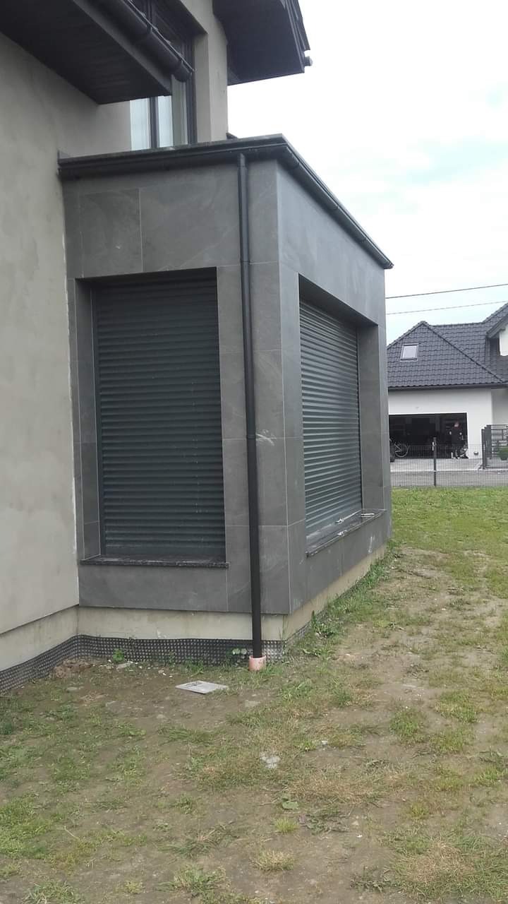 Szary narożny wykusz z zamkniętymi roletami zewnętrznymi, obłożony płytkami imitującymi kamień, na tle trawnika i domu.