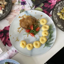 Elegancko podane danie obiadowe na talerzu z marmurkowym wzorem: mięso w sosie własnym, kluski śląskie ułożone w łuk, świeże pomidorki cherry i gałązka koperku, w tle inne talerze z potrawami...