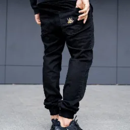 Osoba w czarnych spodniach typu jogger z naszytym złotym logo korony na tylnej kieszeni, czarne sportowe buty z białym logo Nike, szara ściana w tle.