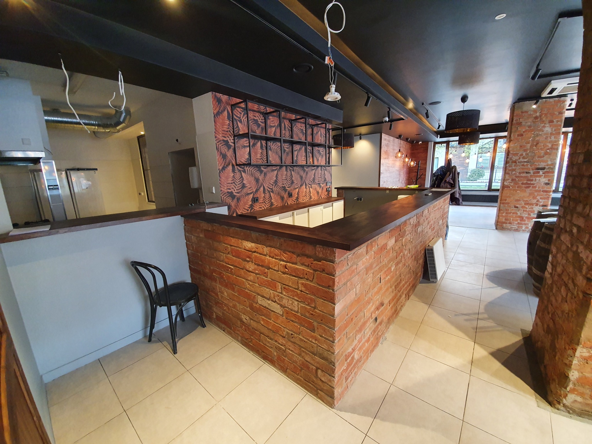 Wnętrze restauracji Endi Wine House w Sopocie: ceglana lada barowa z drewnianym blatem, czarne metalowe regały z motywem liści na ścianie, czarne krzesło i widoczne elementy instalacji sufitowej.