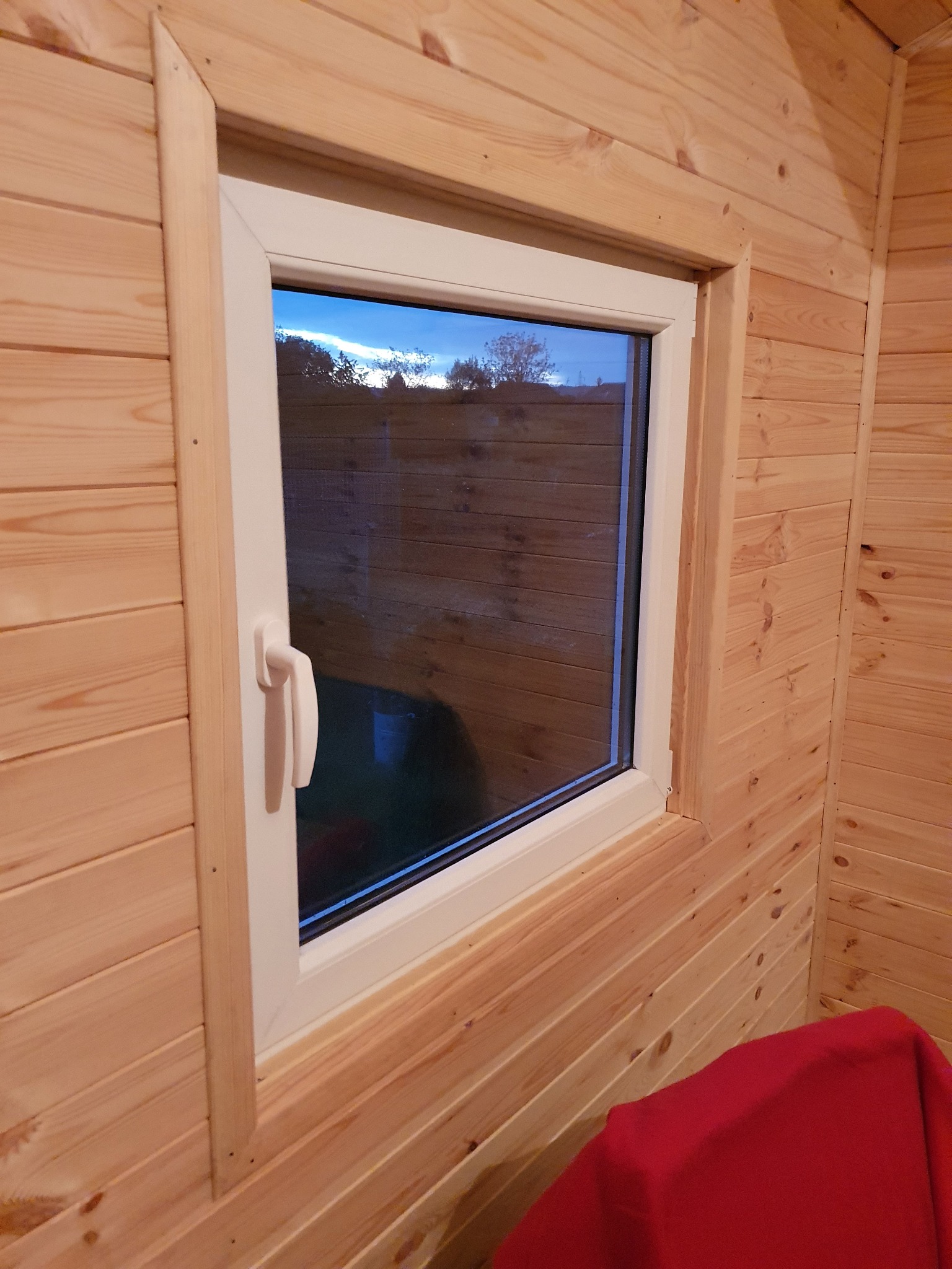 Wnętrze domku letniskowego z widokiem na zewnątrz przez białe okno PCV, ściany wykończone boazerią w naturalnym odcieniu drewna, w tle widoczne drzewa i niebo o zmierzchu.