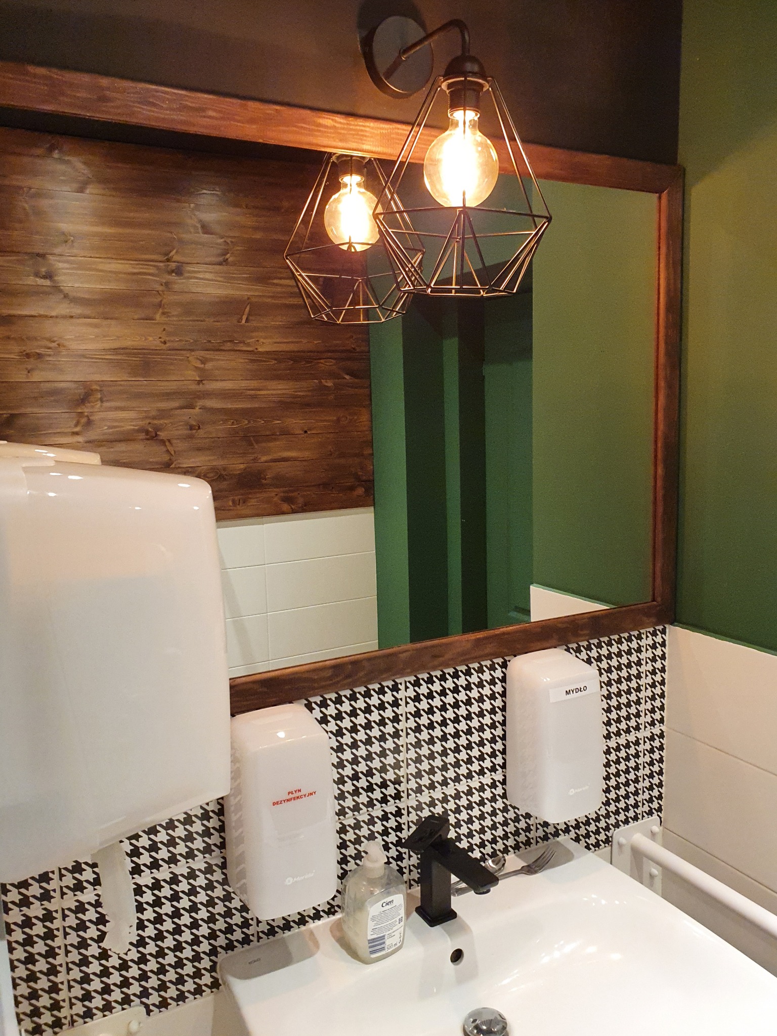 Toaleta w restauracji: umywalka z czarną baterią, dozowniki mydła i płynu dezynfekcyjnego na ścianie w czarno-białą pepitkę, drewniane wykończenie lustra i ściany, nowoczesne lampy z żarówkami...