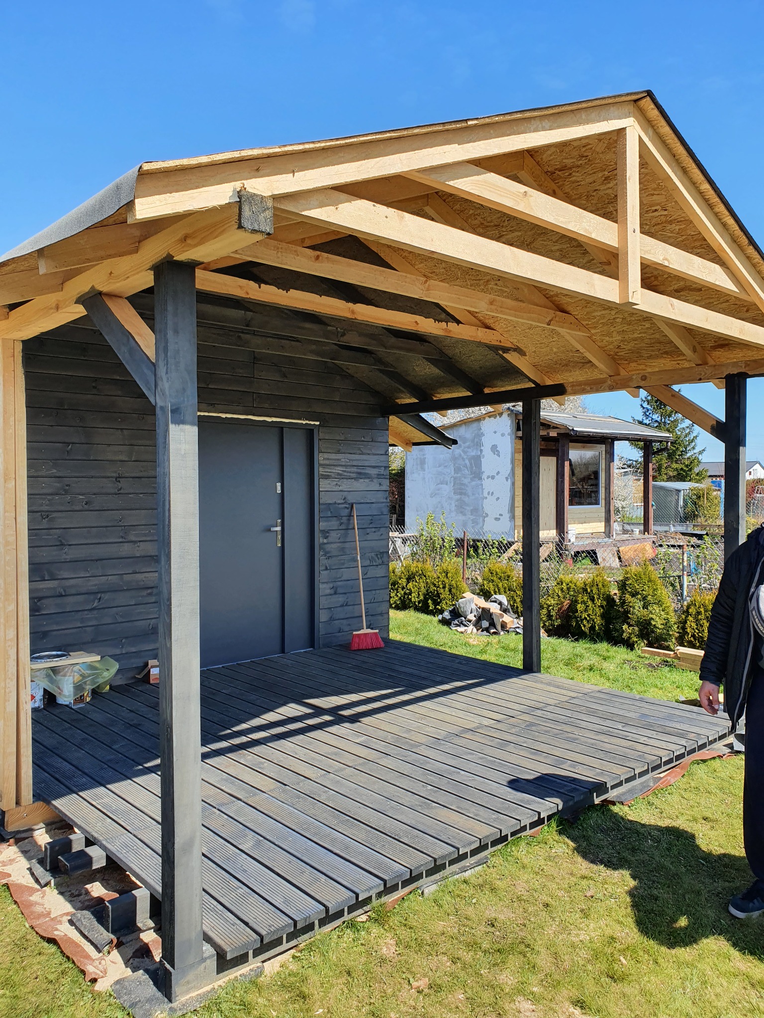 Domek letniskowy w trakcie budowy: szkielet dachu z płyt OSB, czarne drewniane ściany i deski tarasowe, widoczna konstrukcja wsporcza i częściowo ułożona podmurówka.