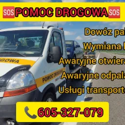 Biała laweta z żółtym pasem i napisem 'POMOC DROGOWA' na poboczu autostrady, przewożąca uszkodzony, granatowy samochód osobowy.