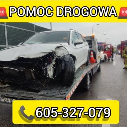 K.D Hol - Uszkodzony biały samochód osobowy na platformie lawety po wypadku drogowym, na mokrej nawierzchni, w tle straż pożarna i ratownicy.