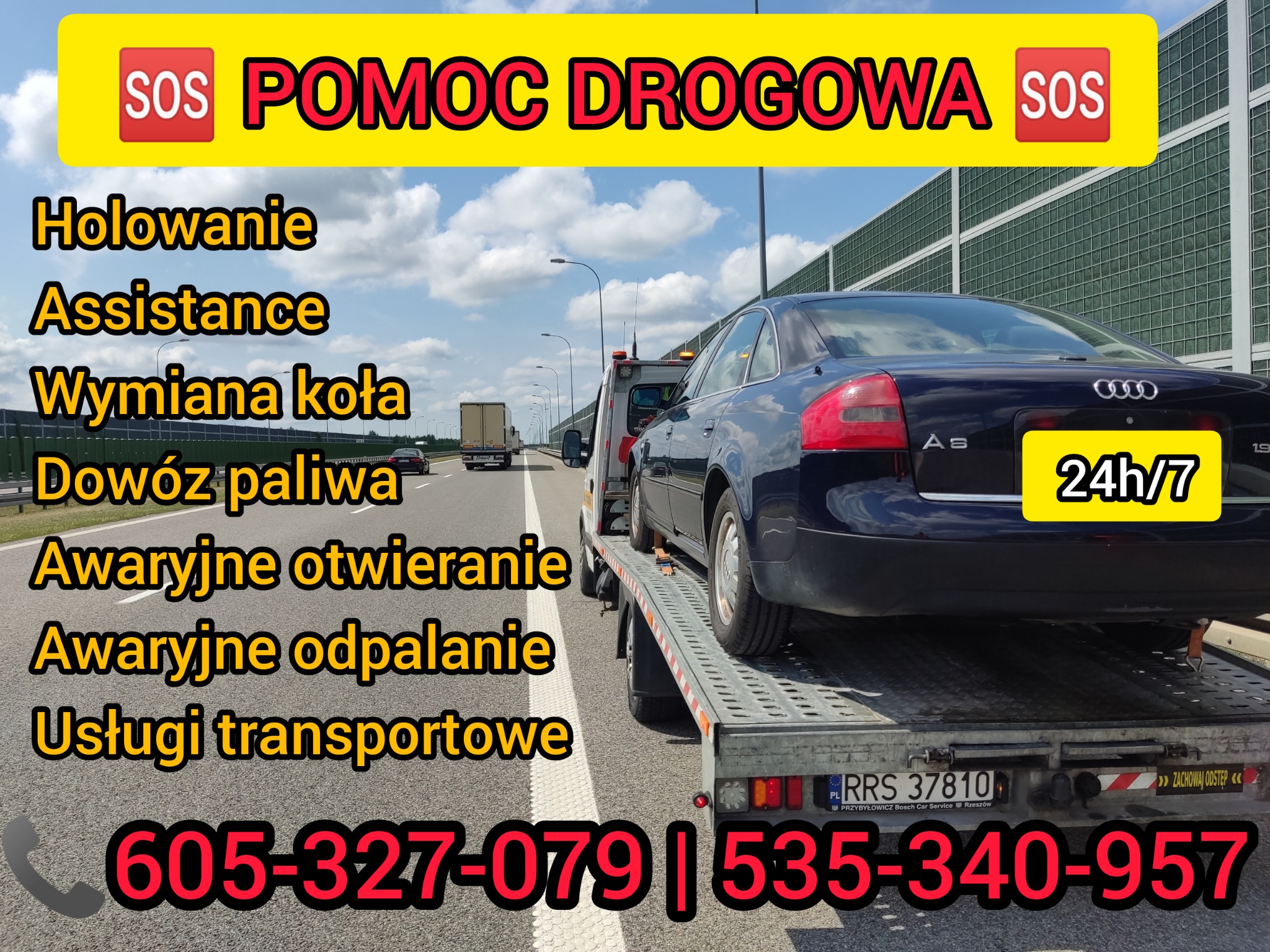Granatowy samochód osobowy marki Audi A6 transportowany na platformie lawety pomocy drogowej na autostradzie, widoczny numer telefonu kontaktowego i hasło '24h/7'.