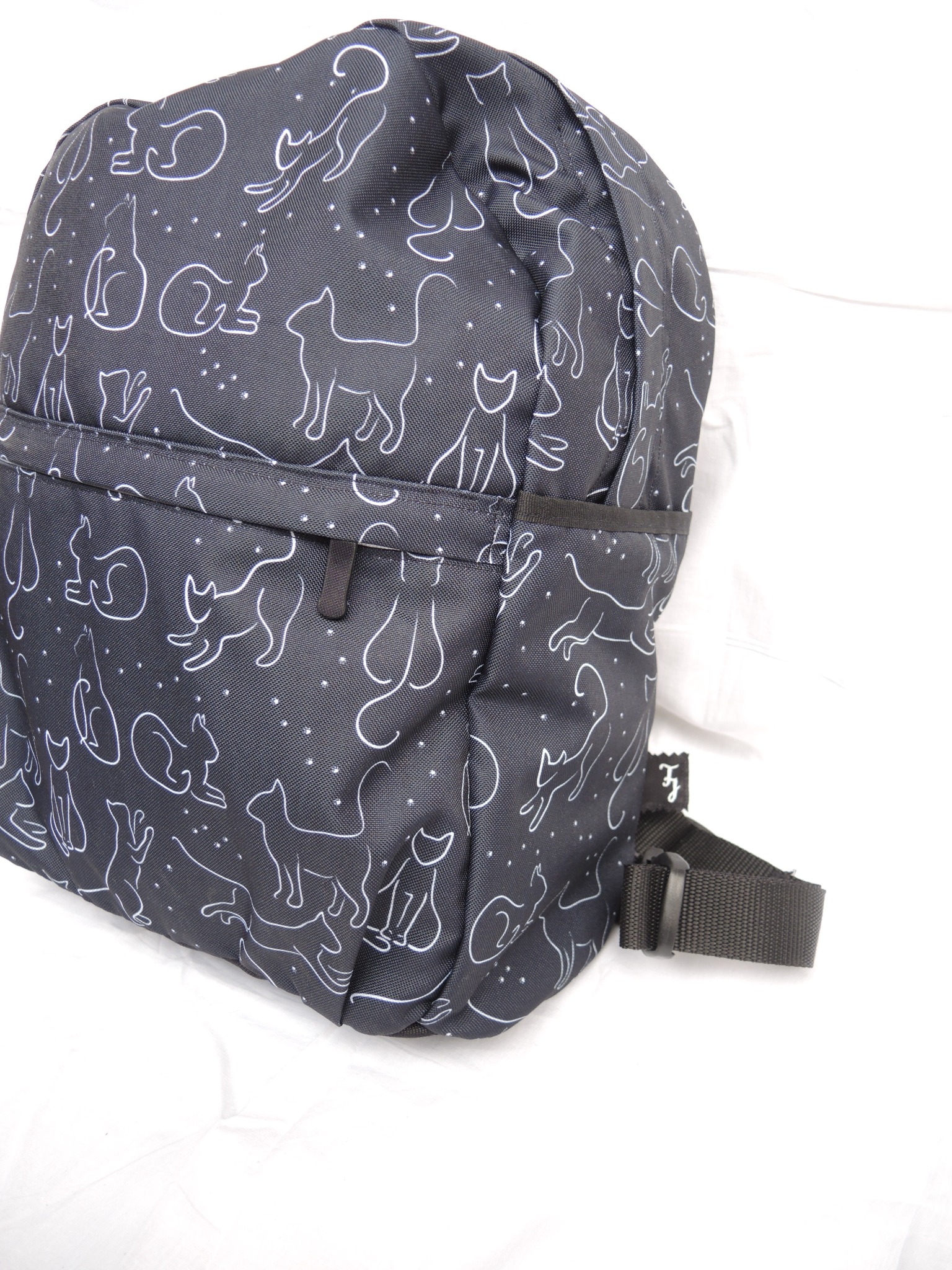 Plecak z czarnego materiału w białe kontury kotów i gwiazdek, widoczny fragment szelki i kieszeń na zamek.
