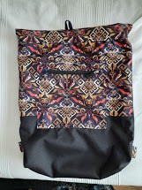 Ręcznie szyty plecak z wzorzystej tkaniny z motywami etnicznymi w odcieniach brązu, czerwieni i czerni, z czarną dolną częścią i kieszenią na zamek błyskawiczny z przodu.