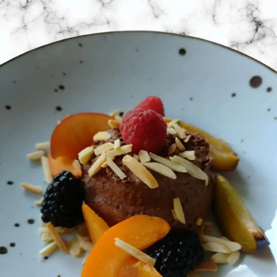 Deser czekoladowy udekorowany malinami, jeżynami, morelami i płatkami migdałów, podany na talerzu z marmurkowym tłem.