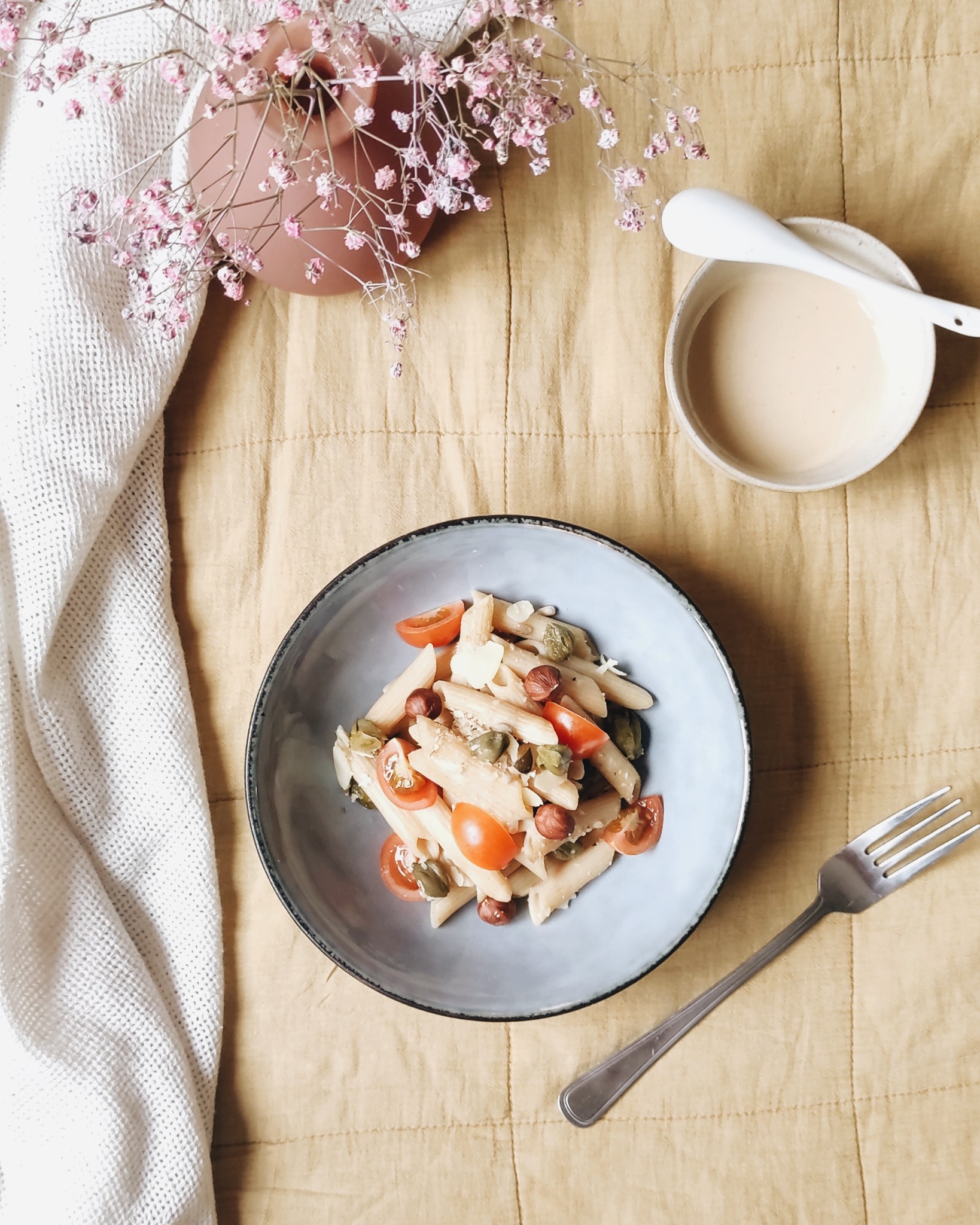 Pastelowa kompozycja z talerzem penne z pomidorkami cherry, orzechami laskowymi i kaparami, miseczką z sosem i bukietem gipsówki na teksturowanym, żółtym tle.