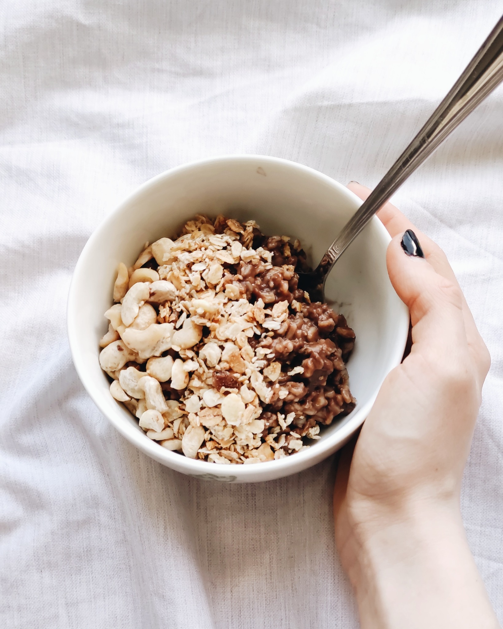 Biała miseczka z owsianką czekoladową i orzechami nerkowca, trzymana w dłoni na tle białej tkaniny.