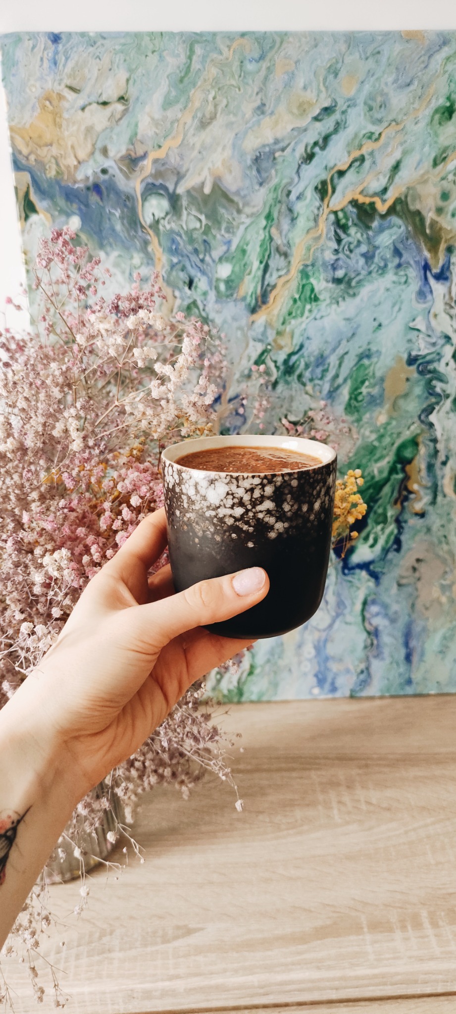 Dłoń trzymająca czarny kubek z deserem czekoladowym, obok bukiet suszonych kwiatów i abstrakcyjny obraz na drewnianym stole.