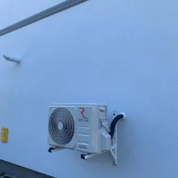 Biała zewnętrzna jednostka klimatyzacji marki Rotenso zamontowana na ścianie budynku, widoczne rury i przewody, żółty znak gazowy z numerem telefonu, tło stanowi biała ściana z fakturą i szara listwa.