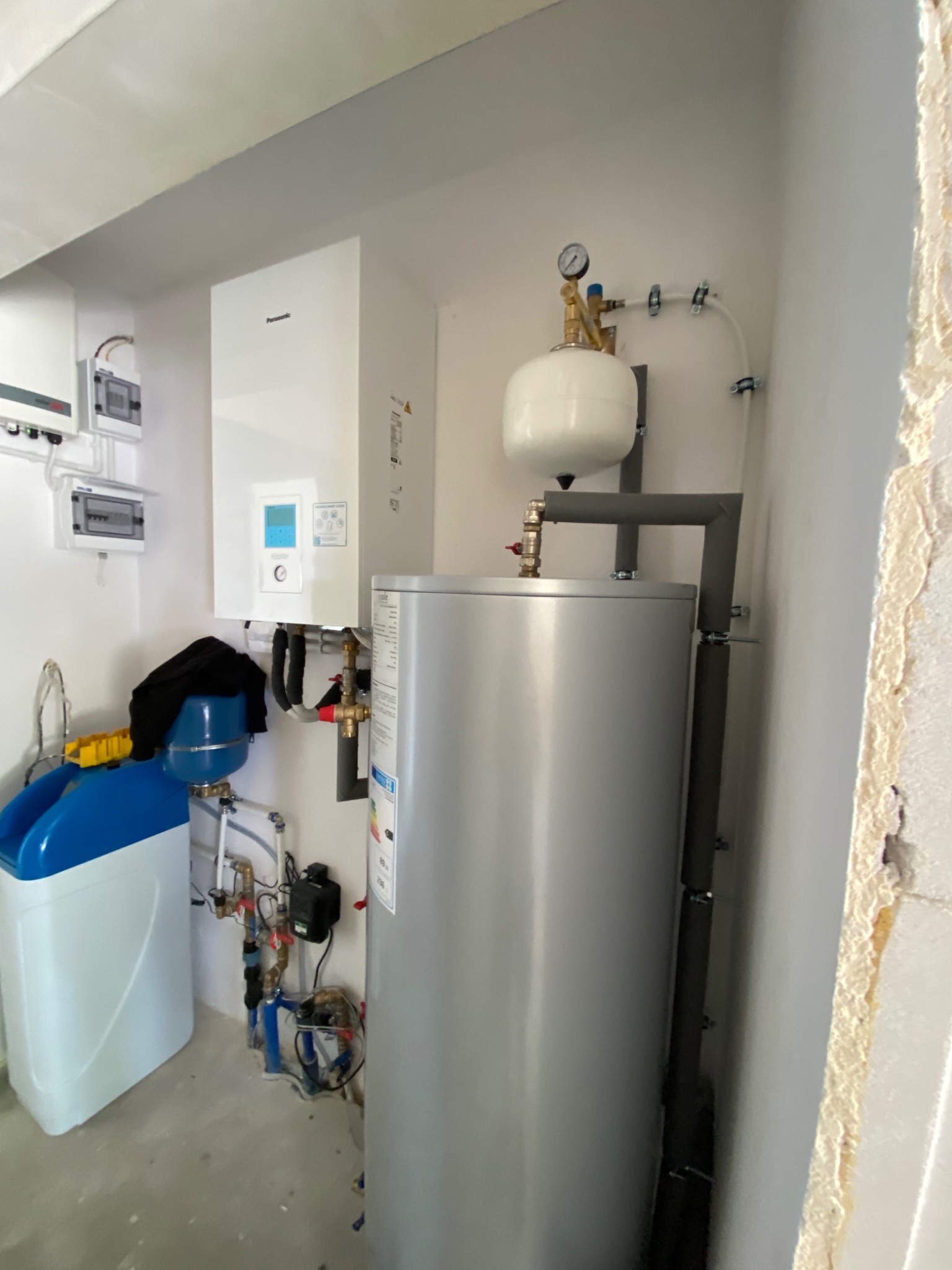Nowoczesny, biały piec gazowy wiszący obok srebrnego zasobnika ciepłej wody z szarą izolacją rur, dodatkowo widoczny zmiękczacz wody i instalacja elektryczna.