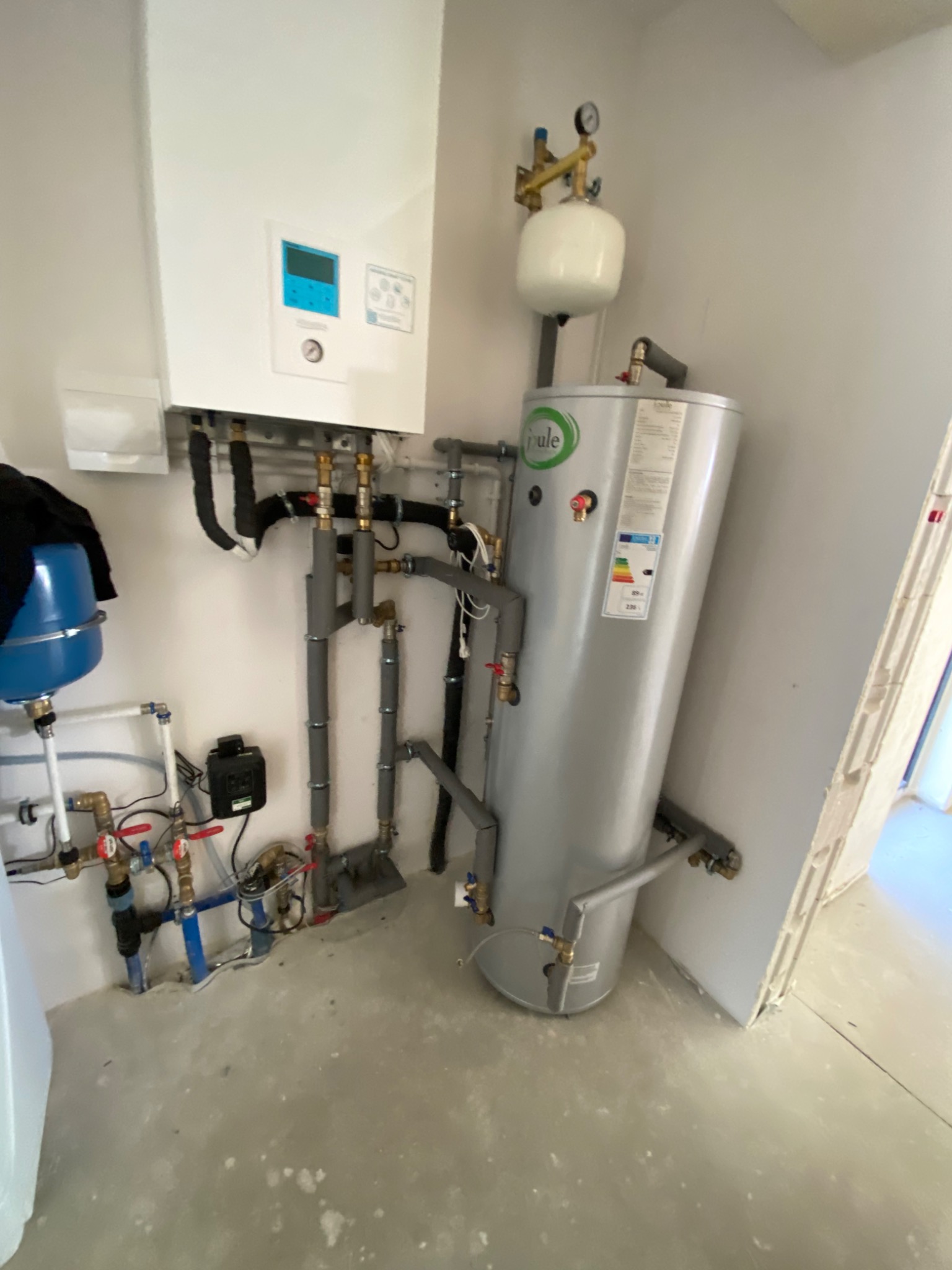 Nowoczesny, kondensacyjny piec gazowy z zasobnikiem ciepłej wody i osprzętem, widoczne rury instalacji grzewczej i hydraulicznej, zamontowany w pomieszczeniu o surowym wykończeniu.