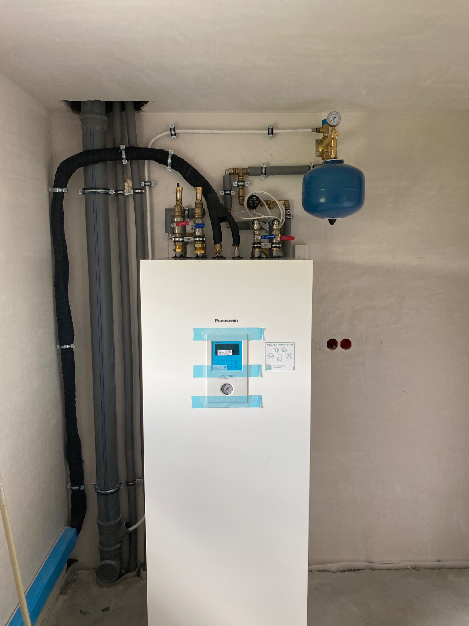 Biała pompa ciepła Panasonic Aquarea Smart Cloud, z niebieskim zbiornikiem wyrównawczym i rurami, zamontowana na surowej ścianie w trakcie instalacji, zabezpieczona taśmą malarską.