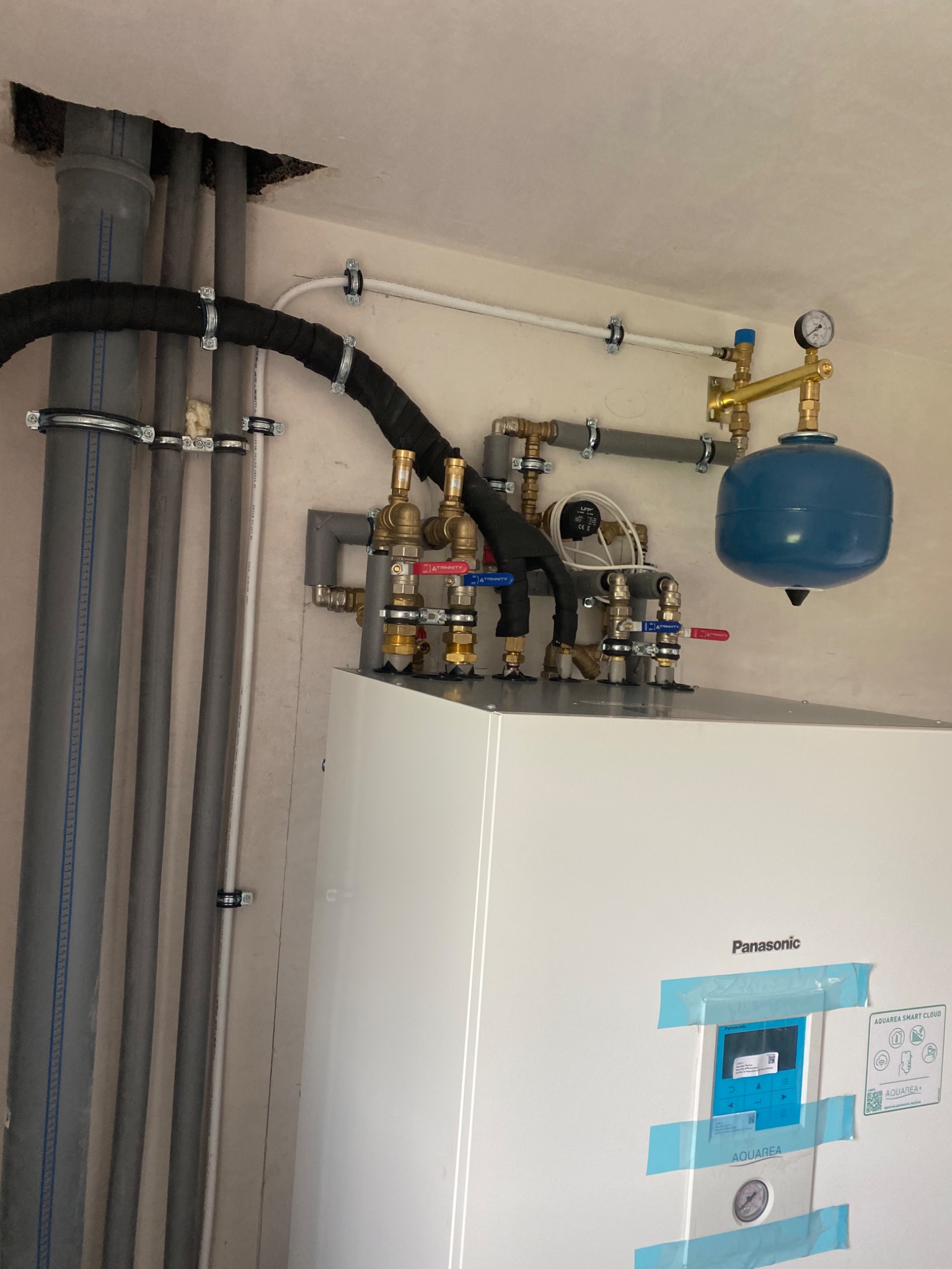 Nowoczesny, biały piec Panasonic Aquarea z niebieskim zbiornikiem wyrównawczym, widoczne rury instalacji grzewczej z zaworami i manometrem, taśma malarska zabezpiecza panel sterowania.
