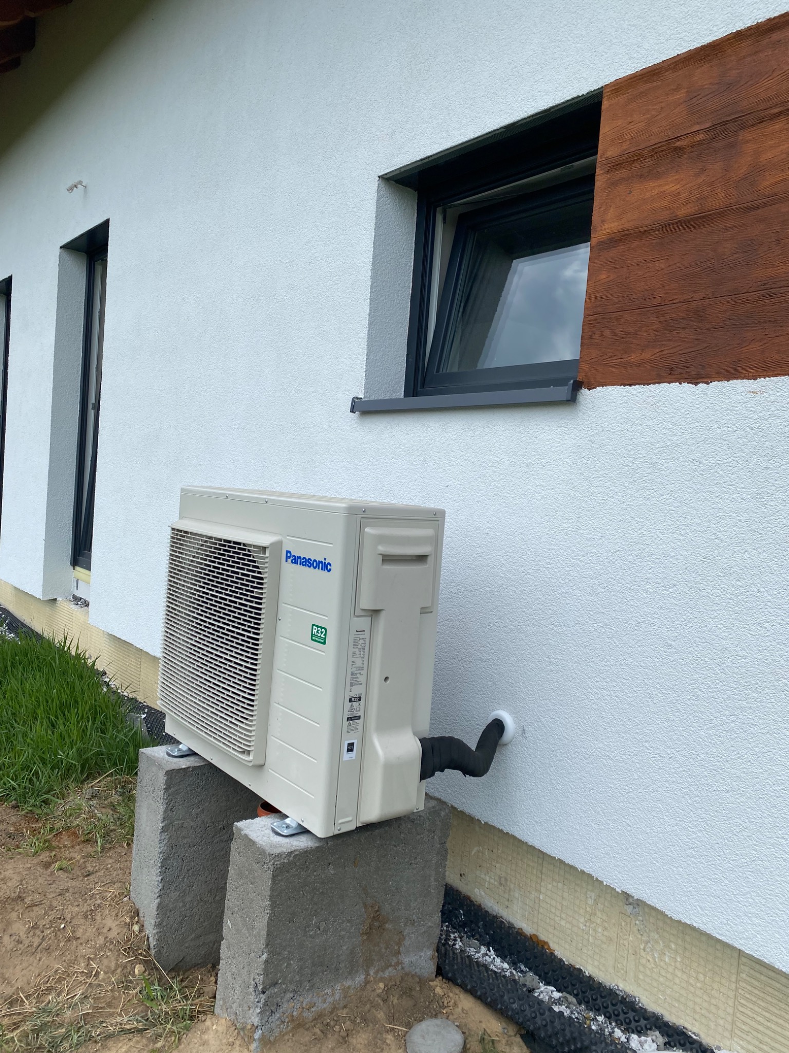 Zewnętrzna jednostka klimatyzacji Panasonic zamontowana na betonowych bloczkach przy białej ścianie budynku z widocznym odpływem skroplin.