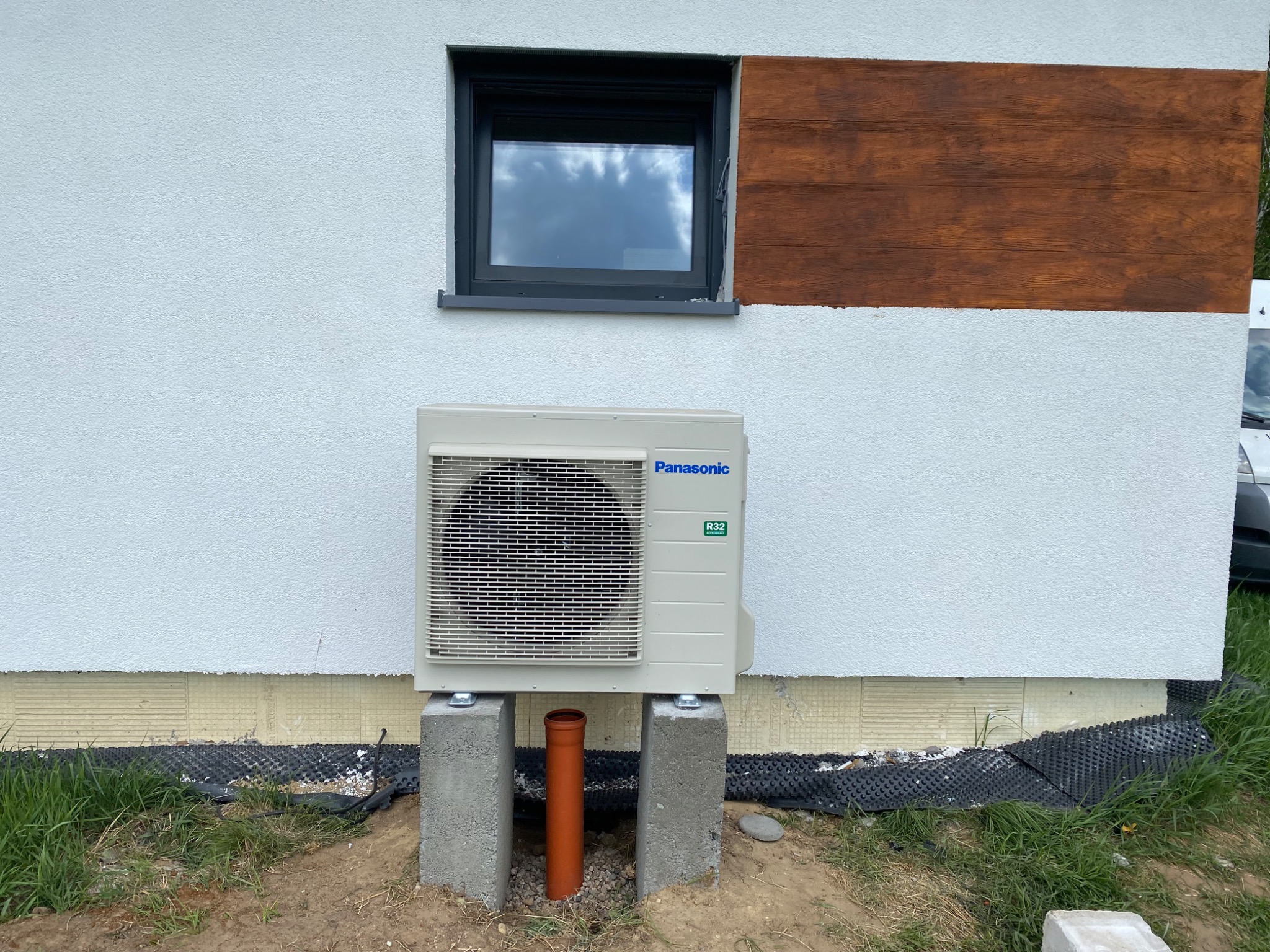 Zewnętrzna jednostka klimatyzacji Panasonic zamontowana na betonowych bloczkach przy ścianie budynku z oknem i drewnianą dekoracją, widoczna pomarańczowa rura odpływowa.