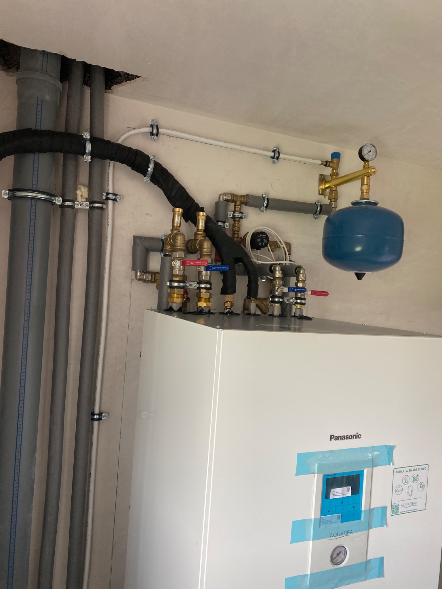 Biały piec Panasonic Aquarea Smart Cloud z niebieskim zbiornikiem wyrównawczym i rurami, świeżo po instalacji, zabezpieczony niebieską taśmą malarską.