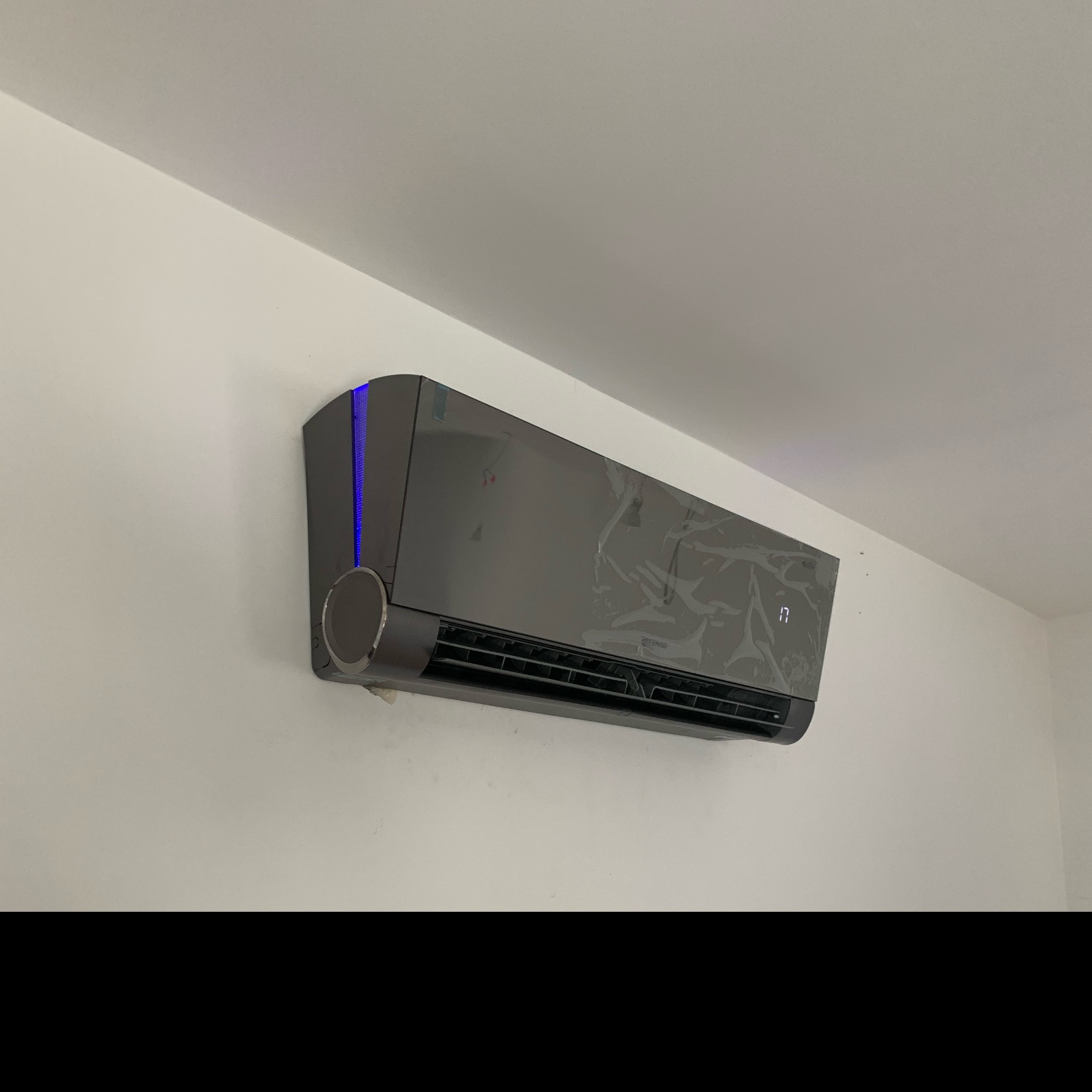 Szary klimatyzator ścienny z niebieskim podświetleniem, częściowo jeszcze w folii ochronnej, zamontowany na białej ścianie.