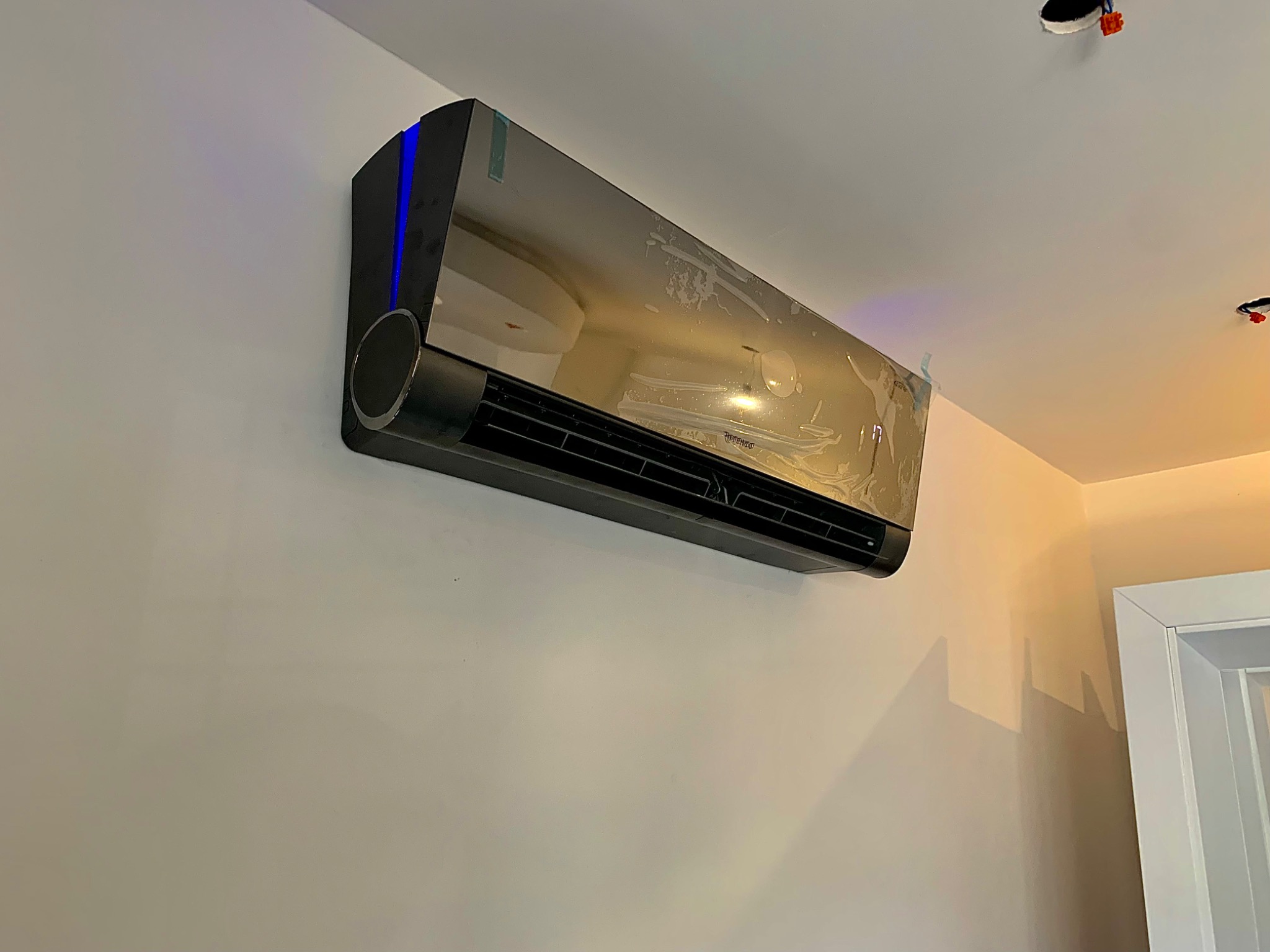 Czarny klimatyzator ścienny z niebieskim podświetleniem LED, wciąż zabezpieczony folią ochronną, zamontowany na beżowej ścianie w nowym wnętrzu.