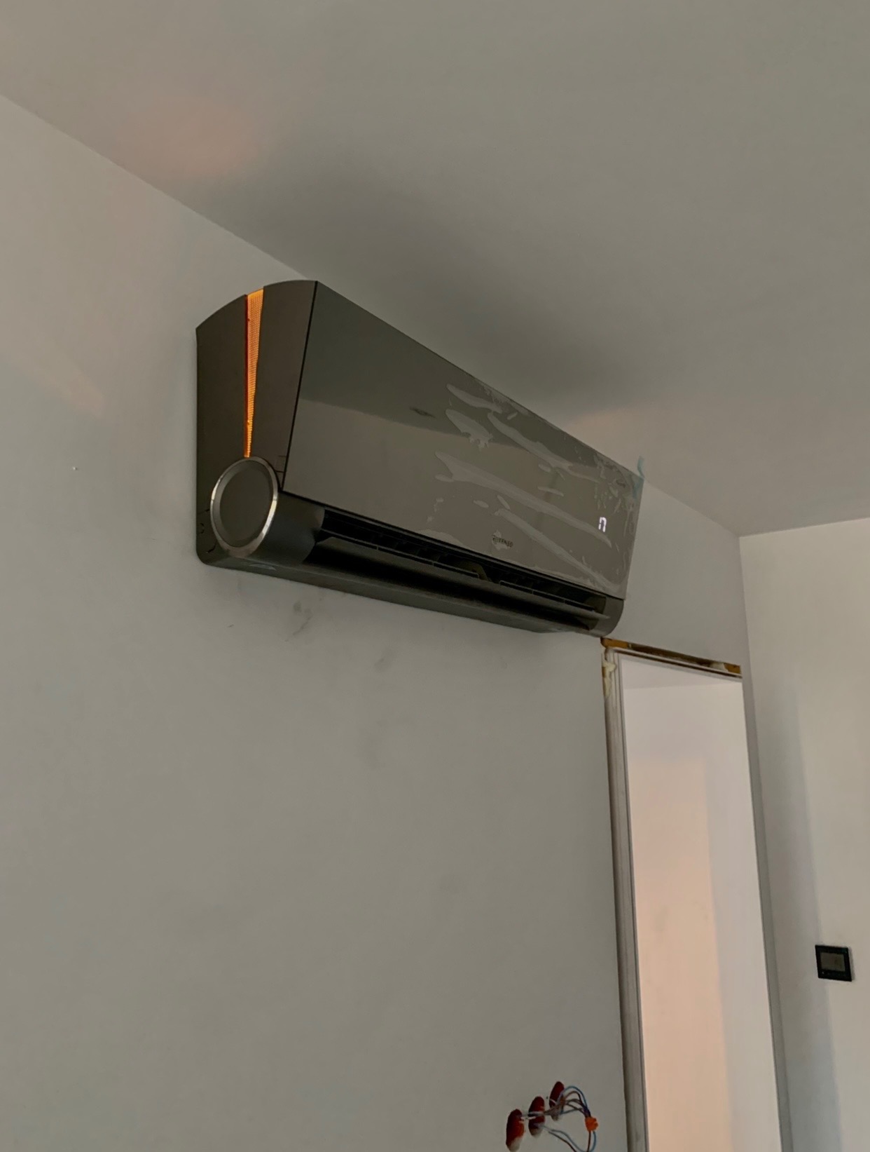 Czarny klimatyzator ścienny zamontowany na szarej ścianie, jeszcze z folią ochronną, widoczne niedokończone okablowanie elektryczne w ścianie.