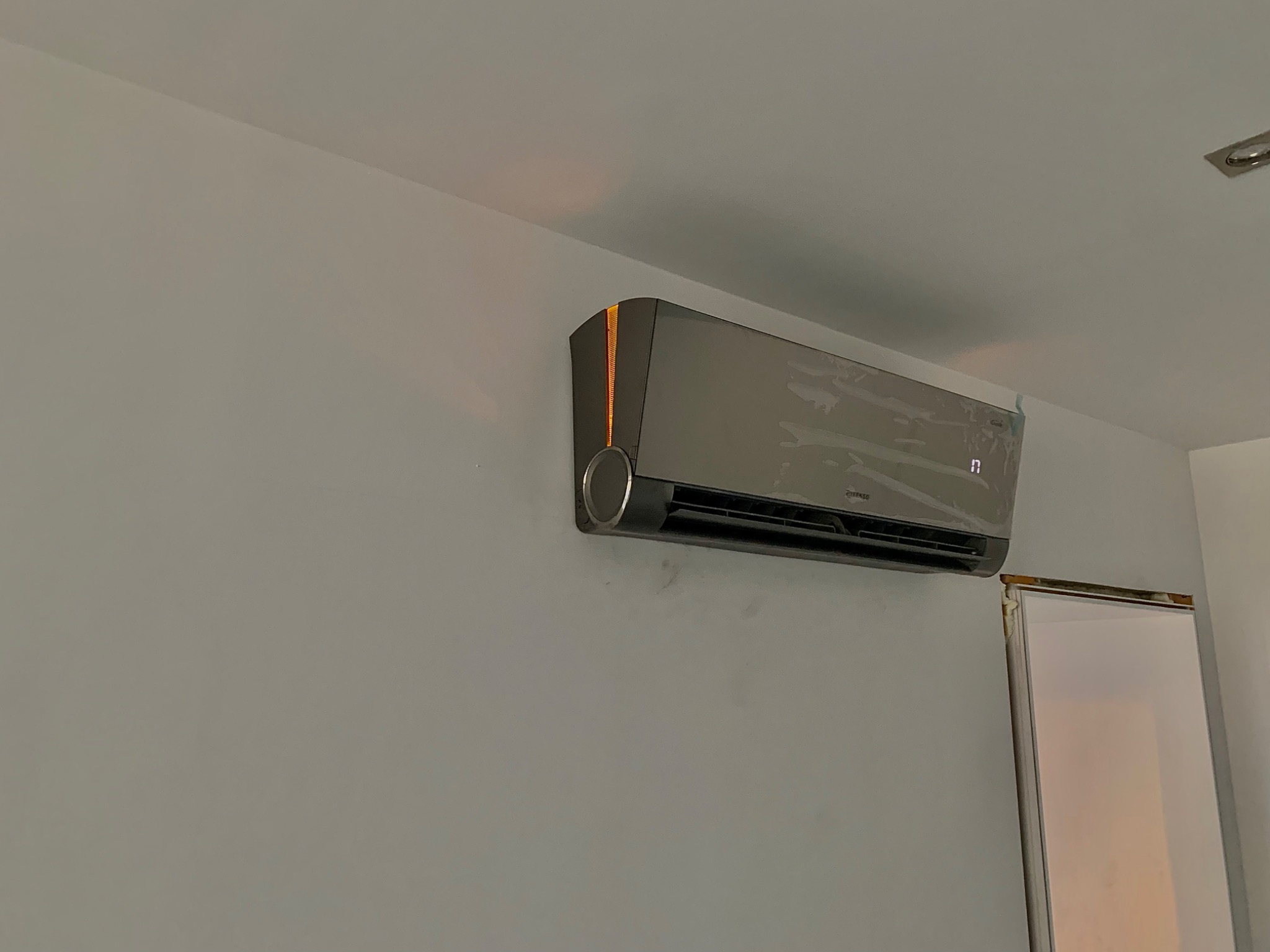 Szary klimatyzator z pomarańczowym akcentem, zamontowany na szarej ścianie, częściowo owinięty folią ochronną, widoczny fragment ościeżnicy drzwi.