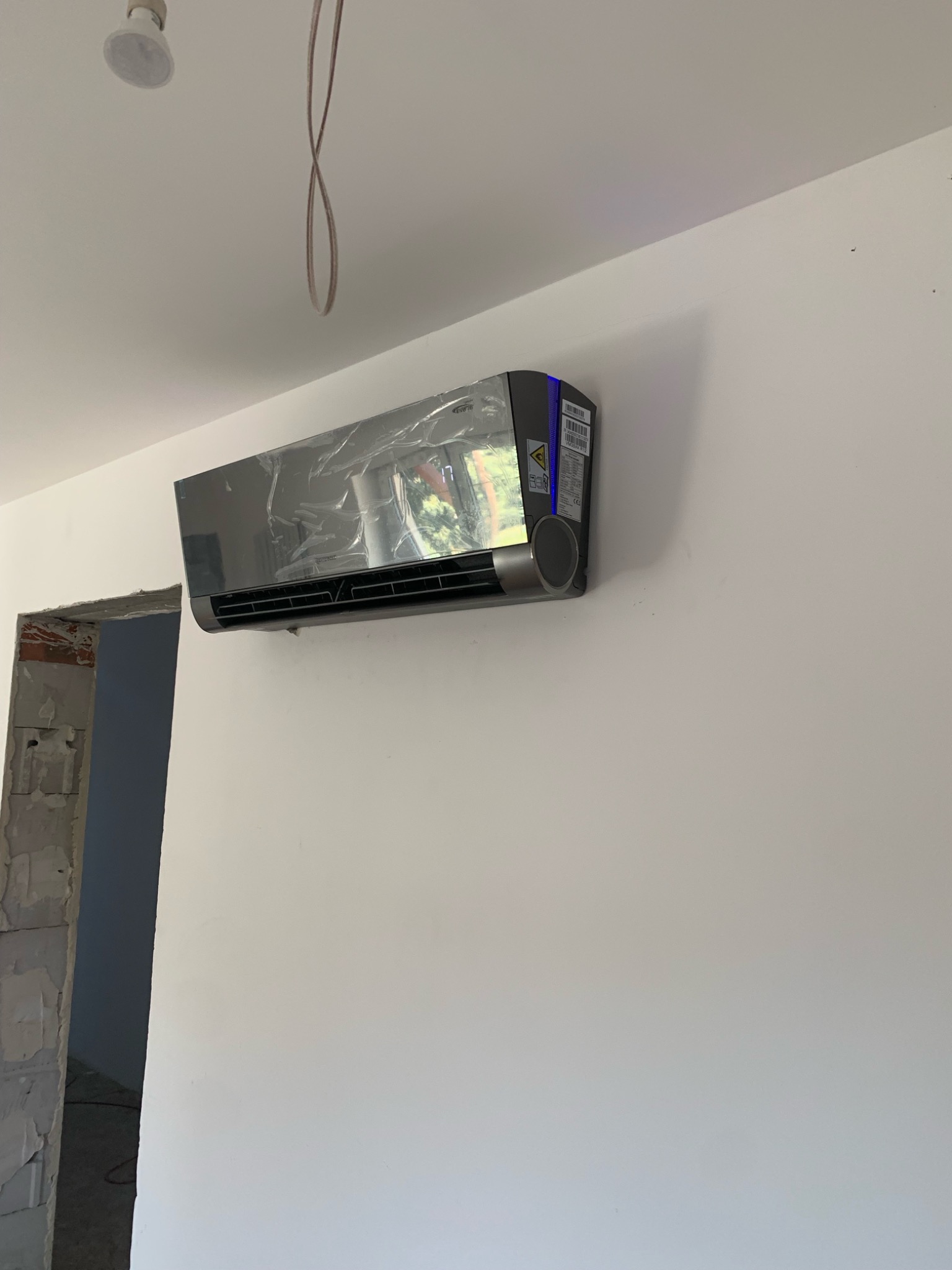 Nowoczesny, srebrny klimatyzator ścienny, jeszcze w folii ochronnej, zamontowany na białej ścianie w nowym budynku, widoczny surowy otwór drzwiowy po lewej stronie.