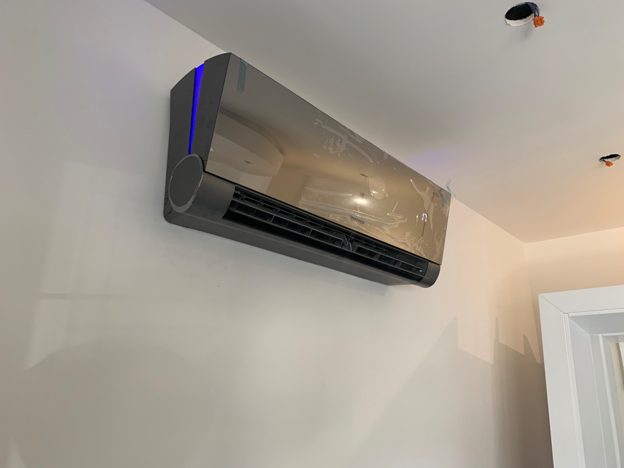 Nowoczesny, ciemnoszary klimatyzator z niebieskim podświetleniem, zamontowany na białej ścianie, z folią ochronną, widoczne otwory w suficie z przewodami.