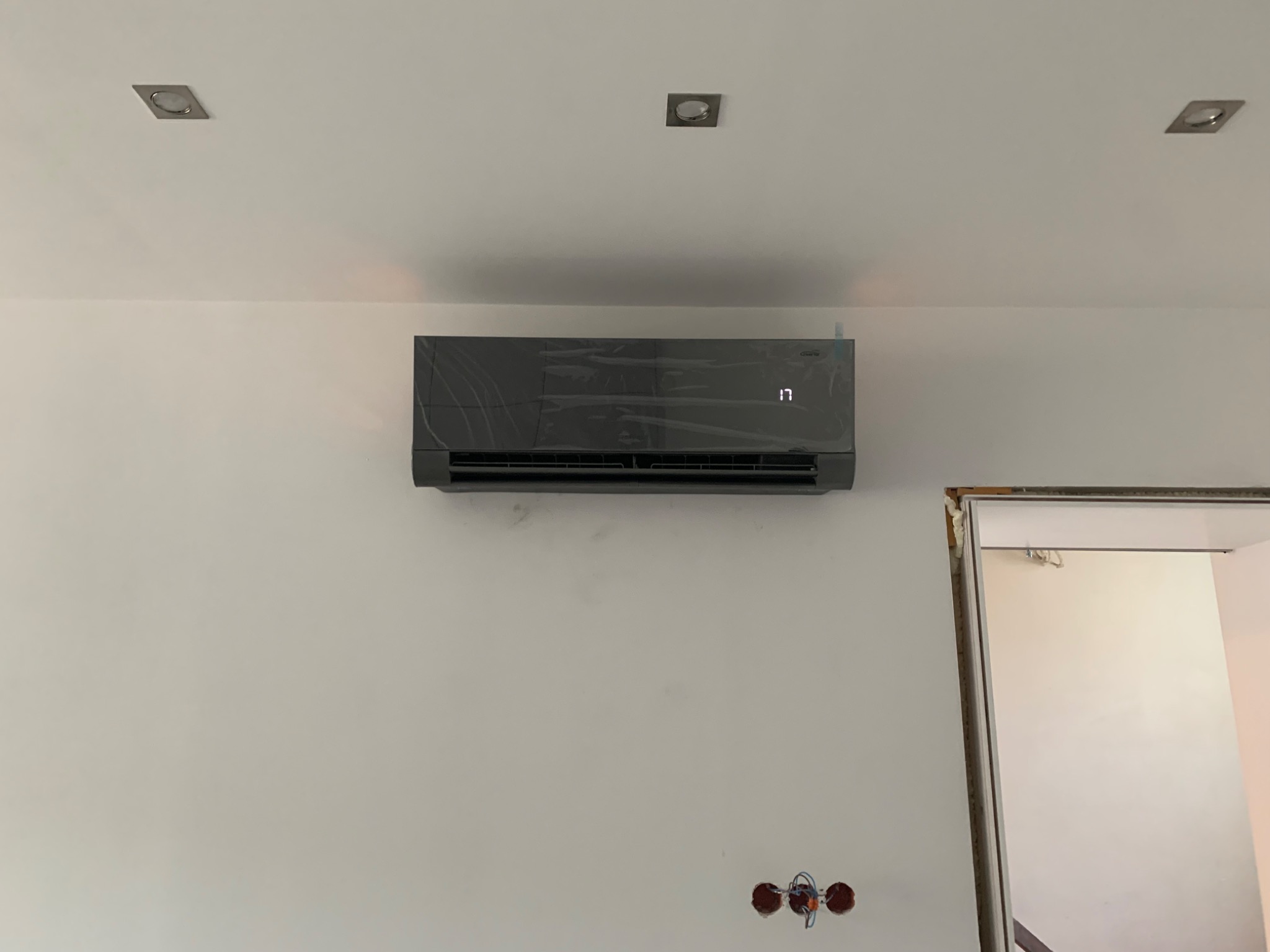 Czarny klimatyzator ścienny, jeszcze w folii ochronnej, zamontowany na białej ścianie, widoczne otwory na kable elektryczne i fragment ościeżnicy drzwi.