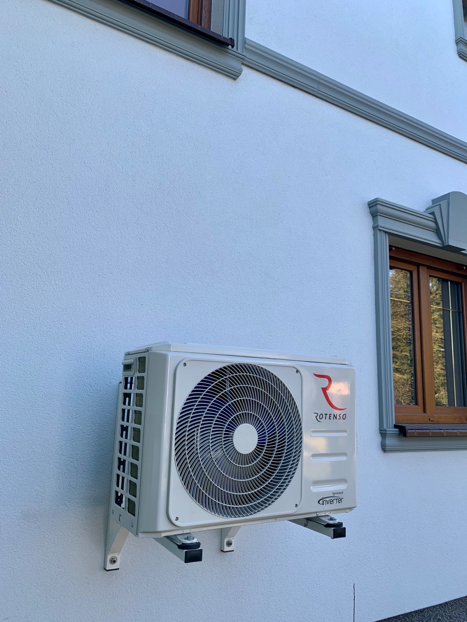 Biała jednostka zewnętrzna klimatyzacji marki Rotenso zamontowana na białej, teksturowanej ścianie budynku z ozdobnymi elementami okiennymi.