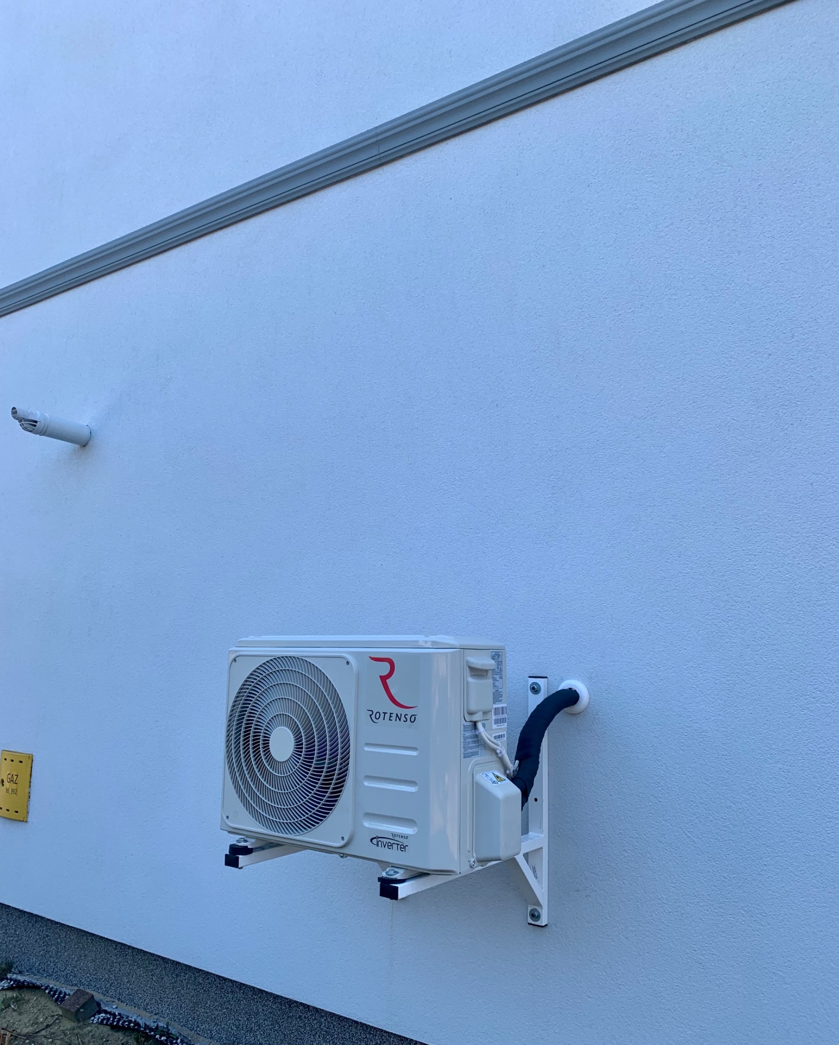 Zamontowana jednostka zewnętrzna klimatyzacji Rotenso na białej ścianie budynku, widoczne rury w izolacji i elementy mocujące.