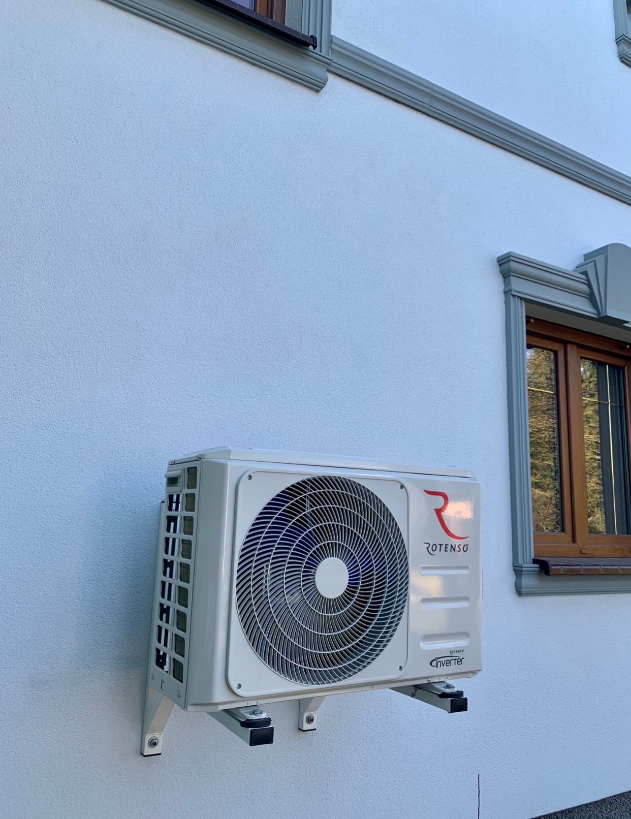 Biała jednostka zewnętrzna klimatyzacji Rotenso zamontowana na ścianie budynku z oknem w drewnianej ramie.
