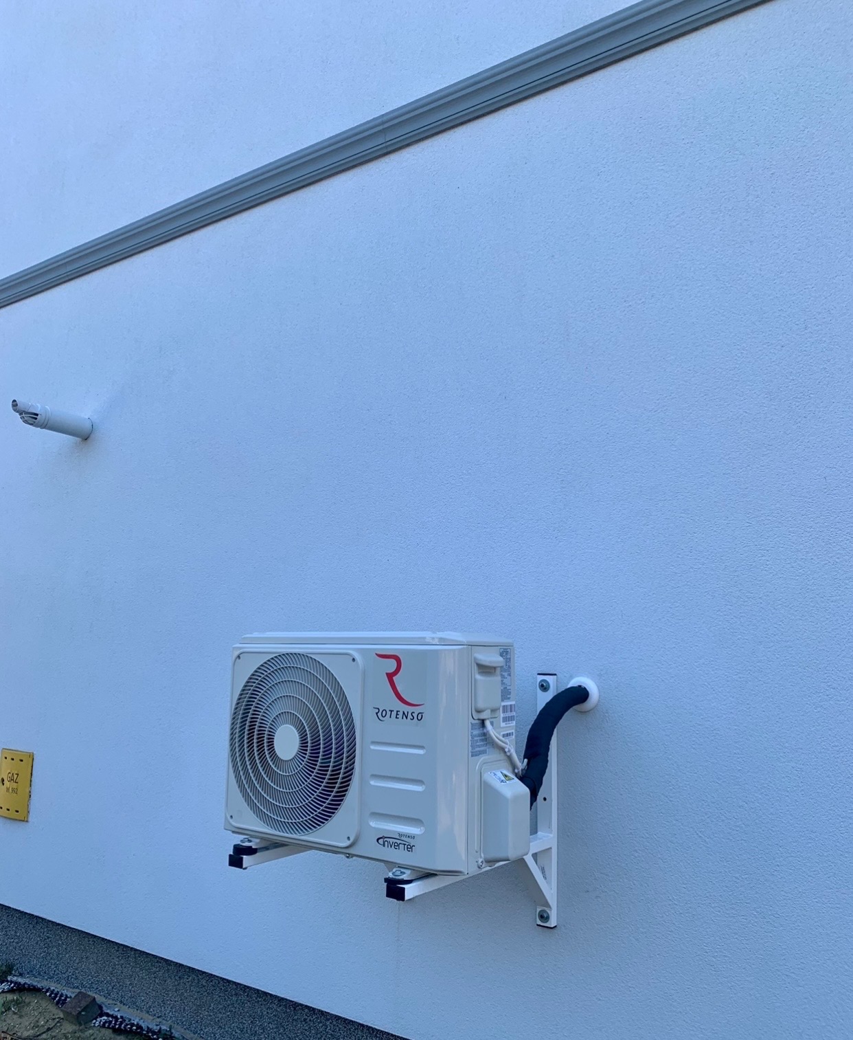 Biała zewnętrzna jednostka klimatyzacji Rotenso zamontowana na białej ścianie budynku, widoczna rura odprowadzająca skropliny i tabliczka gazowa z boku.