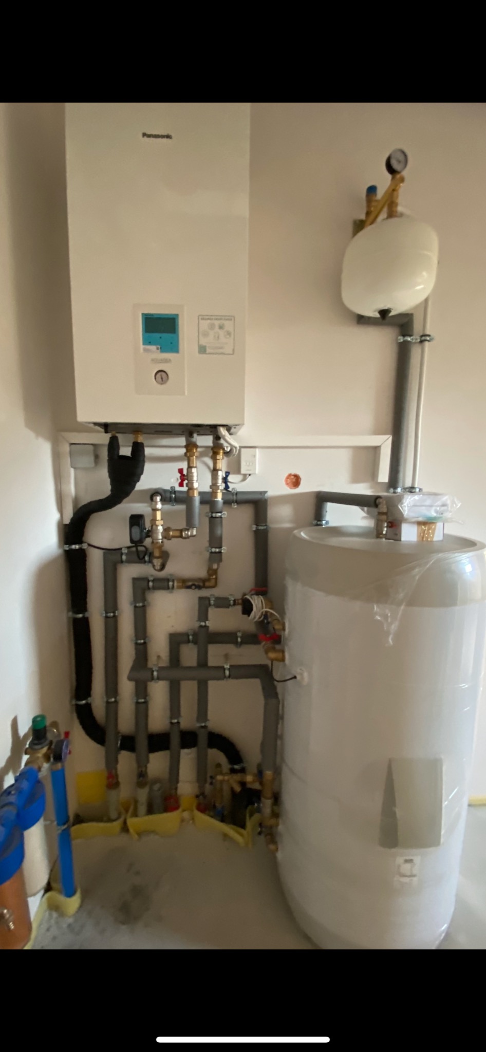 Biały piec gazowy Panasonic zamontowany na ścianie, z widocznymi rurami, zaworami i zbiornikiem wyrównawczym. Nowa instalacja grzewcza z elementami jeszcze w folii ochronnej.