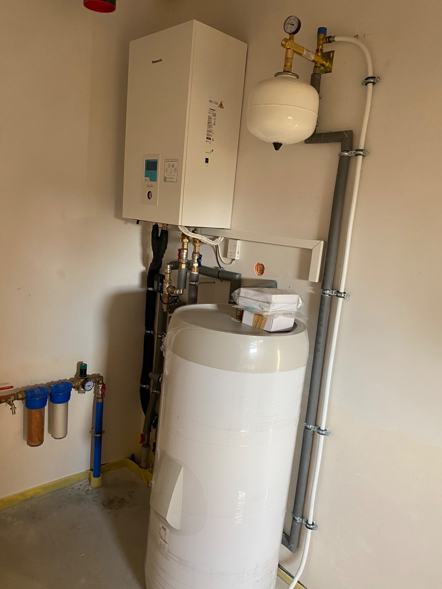 Biały piec CO marki Panasonic zamontowany na ścianie, obok biały zbiornik na wodę owinięty folią, widoczne rury i zawory, instalacja hydrauliczna w pomieszczeniu.
