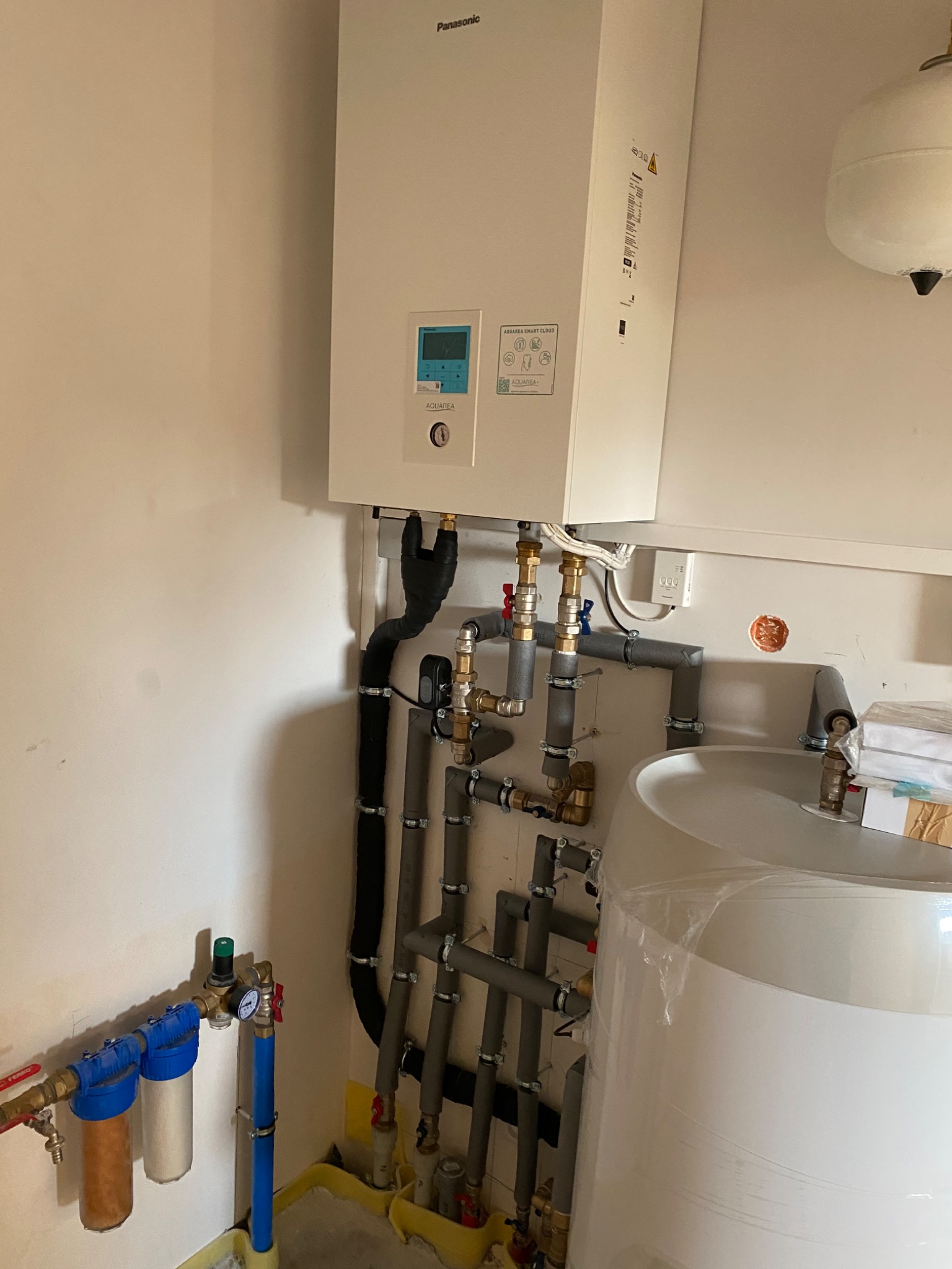 Biały piec Panasonic Aquarea z podłączonymi rurami, filtrami i zbiornikiem, widoczne liczne złączki, zawory i manometr.